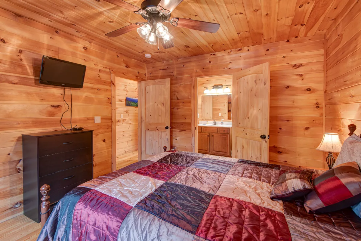 A cozy bedroom with a bed covered in a colorful quilt, a wooden dresser, and a TV mounted on the wall. Double doors open directly into an ensuite bathroom, and warm wood tones envelop the space, giving it a welcoming, rustic cabin feel.