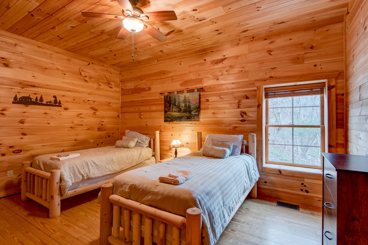 A tranquil bedroom scene featuring wooden beds, a lamp on the bedside table, and a painting on the wall. Natural light streams in, creating a relaxing atmosphere suitable for both adults and children.