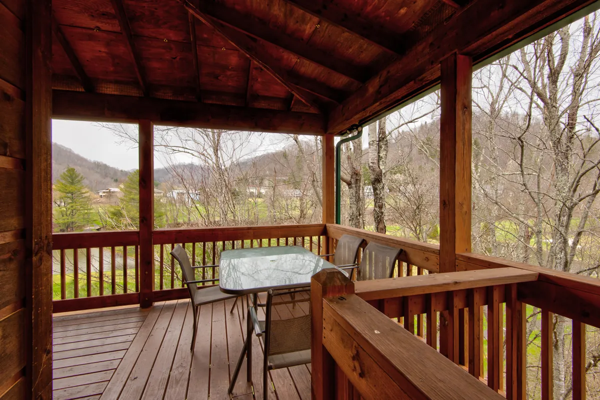A wooden deck features a glass dining table and several empty chairs, all facing a beautiful mountain view. The scene is set for alfresco dining, with the serene landscape in the background, perfect for enjoying meals outdoors while taking in the vistas.
