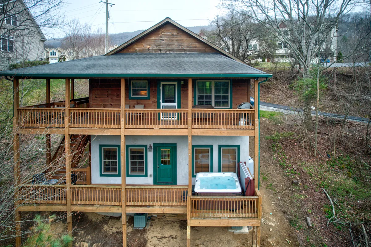This image depicts a welcoming exterior view of the home's wooden deck, featuring a hot tub, green doors, and expansive windows. Surrounded by trees, the deck offers space for outdoor gatherings and relaxation with beautiful nature views.