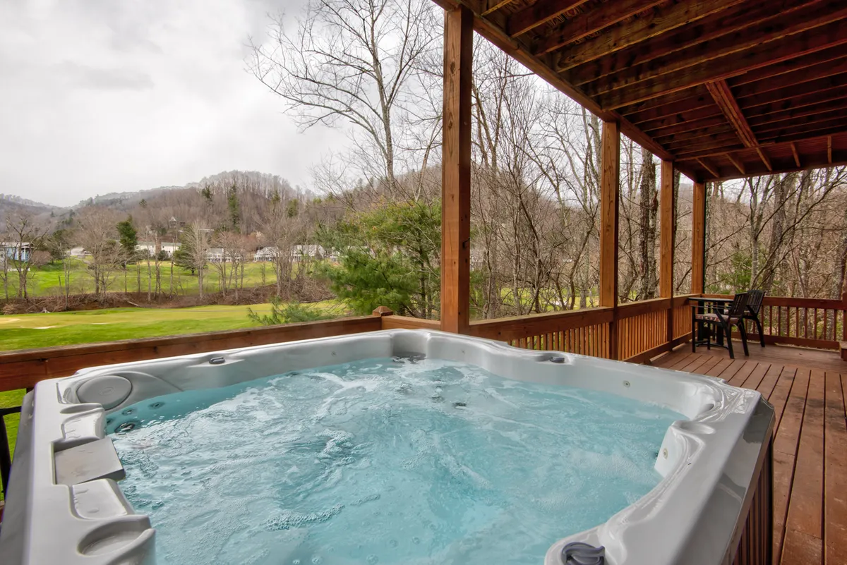 This exterior image features a hot tub on a wooden deck surrounded by trees, with sunset light providing a tranquil backdrop. The deck offers a panoramic view of the mountains, and the hot tub setting invites relaxation and soaking while enjoying nature. The scene captures the peacefulness and privacy of the retreat.