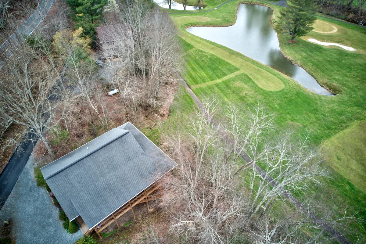 A wide view that features the house set beside a tranquil pond, with an open expanse of green fairway and the adjacent golf course visible. The scene includes golfers in the distance, serene water reflections, and lush natural landscaping, all framed by mountains.