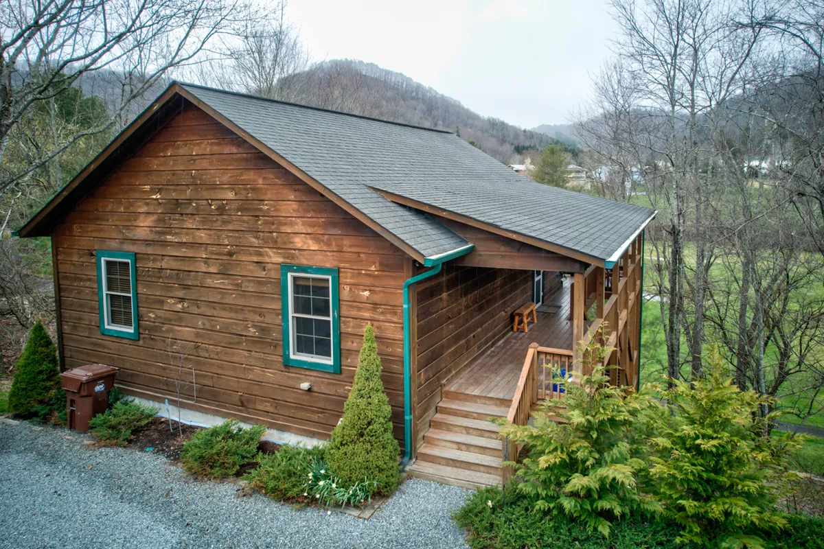 A charming wooden cabin with green trim sits nestled among mature trees, with a welcoming front porch that invites guests to relax and take in the natural surroundings. The exterior looks spacious, with clear areas around the cabin ideal for outdoor activities and for pets to roam safely. The scene radiates an inviting, homey feel perfect for pet owners seeking an immersive nature experience with their dogs.