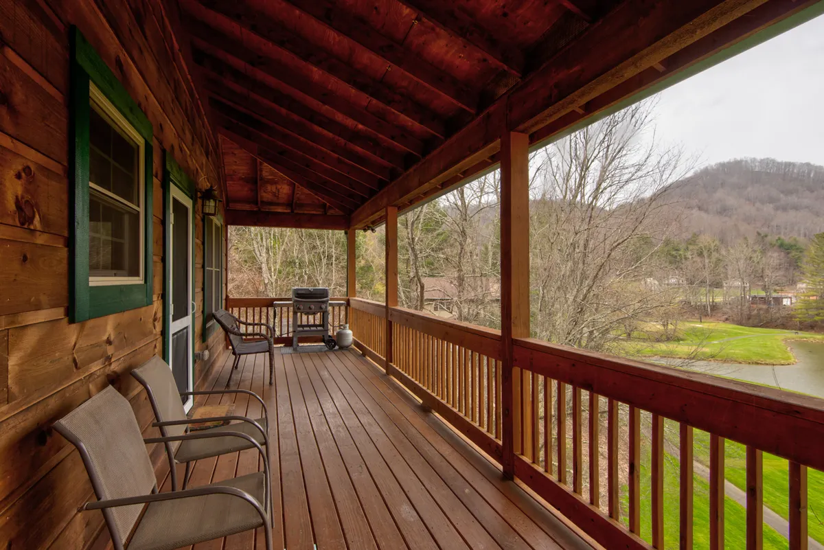 A spacious wooden deck with comfortable seating areas overlooks a lush golf course and distant mountains. A barbecue grill is set up for alfresco cooking, and the setting is surrounded by mature trees, creating a serene and inviting outdoor living space ideal for socializing and relaxation.