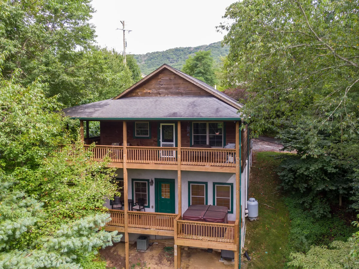The image focuses on a tranquil wooden deck with a green door and a hot tub, surrounded by lush trees. The deck is styled for relaxation and entertaining, capturing sunset views and stargazing potential.