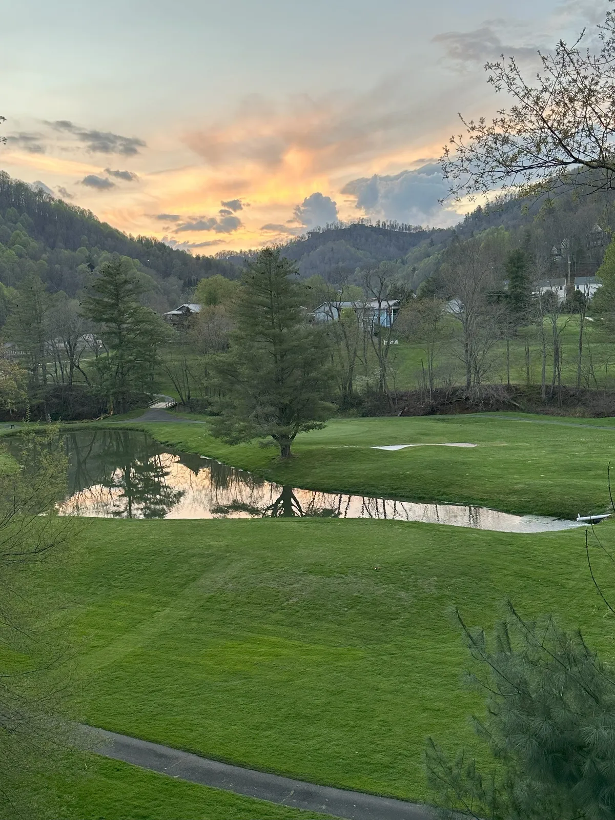A tranquil scene capturing a reflective pond at sunset, bordered by a lush green lawn and framed by distant hills and trees. The expansive sky is filled with soft colors, inviting viewers to imagine peaceful walks, picnics, or photography sessions in the serene landscape.