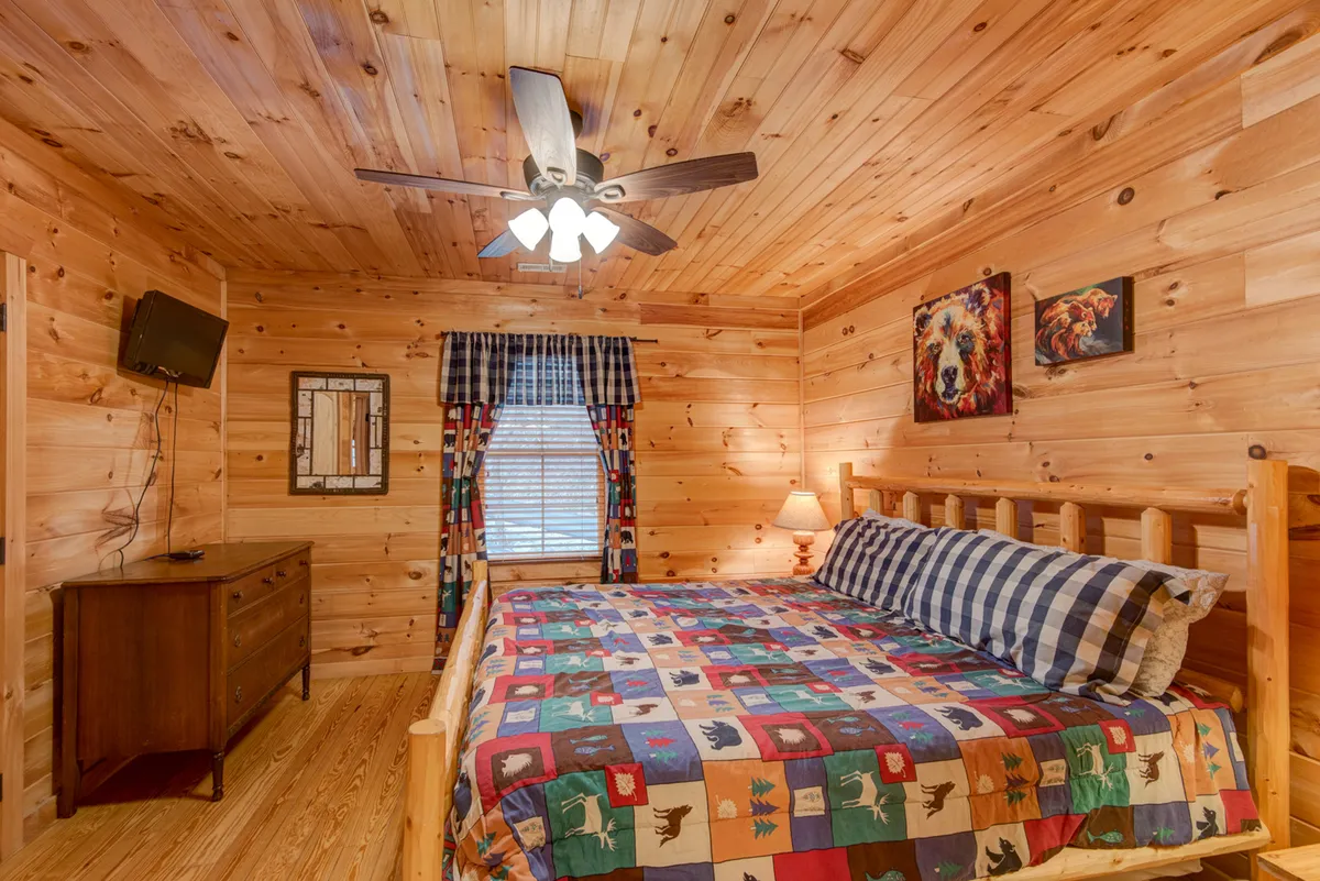 A cozy bedroom with a log cabin-style bed covered by a colorful quilt, a wooden dresser, wall artwork, and a ceiling fan. The space feels rustic and inviting, perfect for relaxing after a day outdoors with pets.