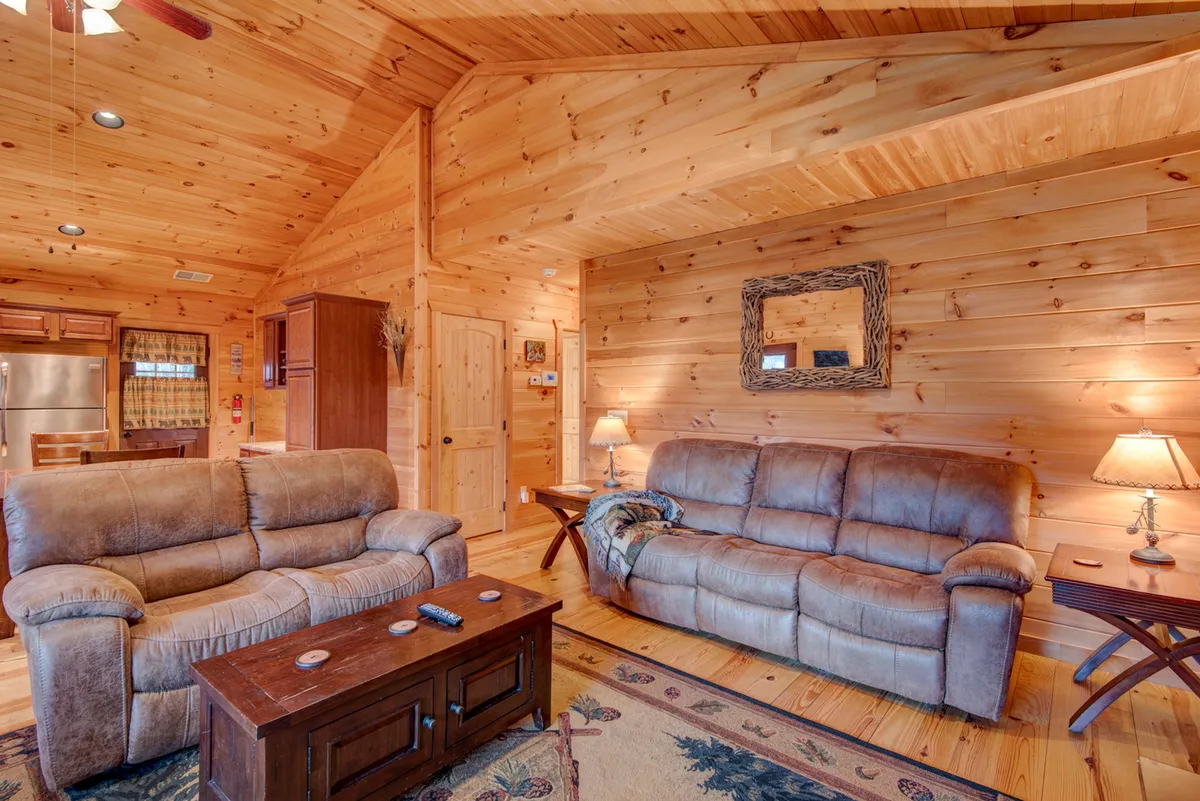 A cozy living room with comfortable sofas, a wooden coffee table, a decorative mirror, and warm lighting from lamps. The space is styled with inviting wood tones and arranged for socializing or movie viewing, making it ideal for family gatherings.