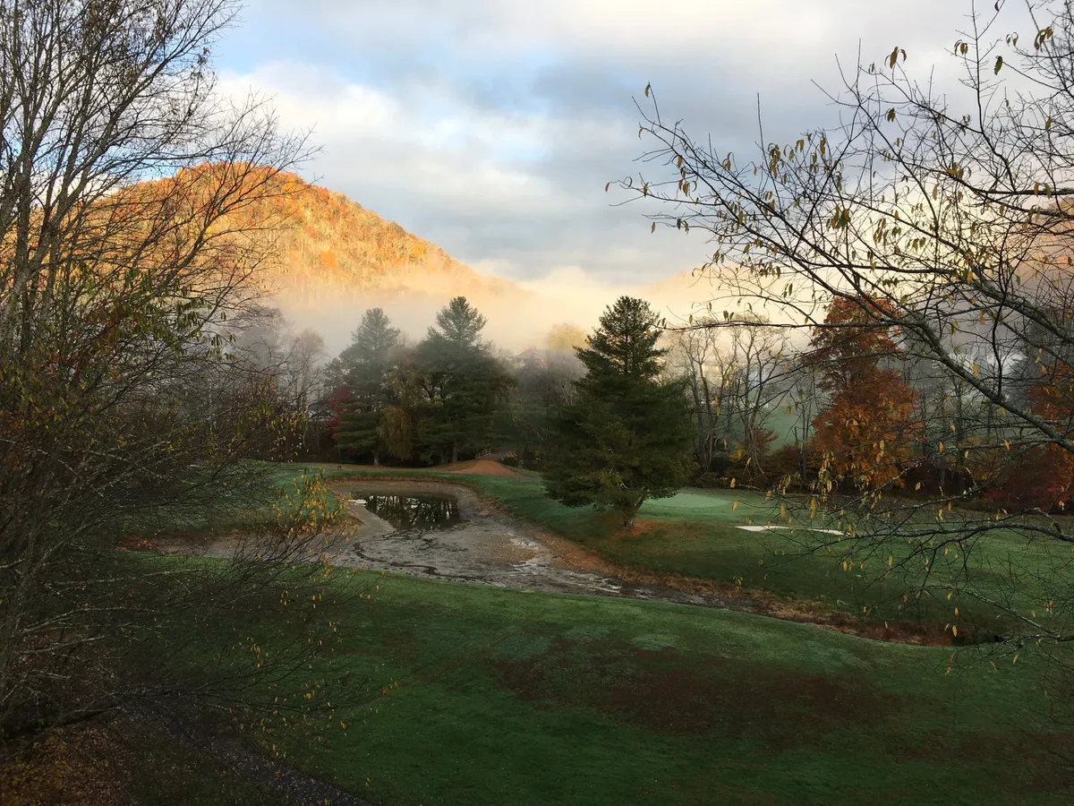 A serene view of a mist-covered mountain at sunrise, lush green grass, a small pond, and trees with autumn-colored leaves. The mist and soft light create a sense of early-morning adventure and connection to the landscape.