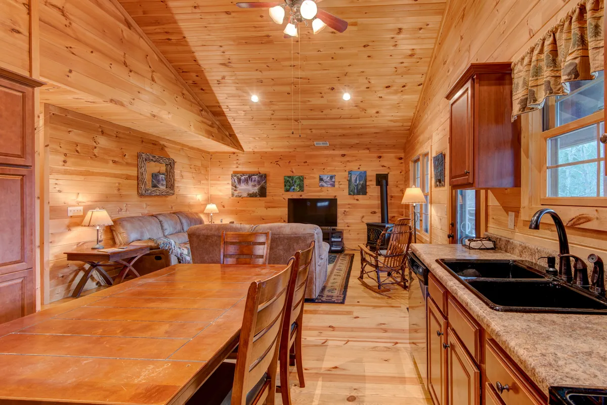 A multifunctional family room featuring a wooden dining table, sofa, television, kitchen sink, and wall art—arranged to support both dining and entertainment. The warm decor and practical layout suggest a space for both relaxing and socializing.