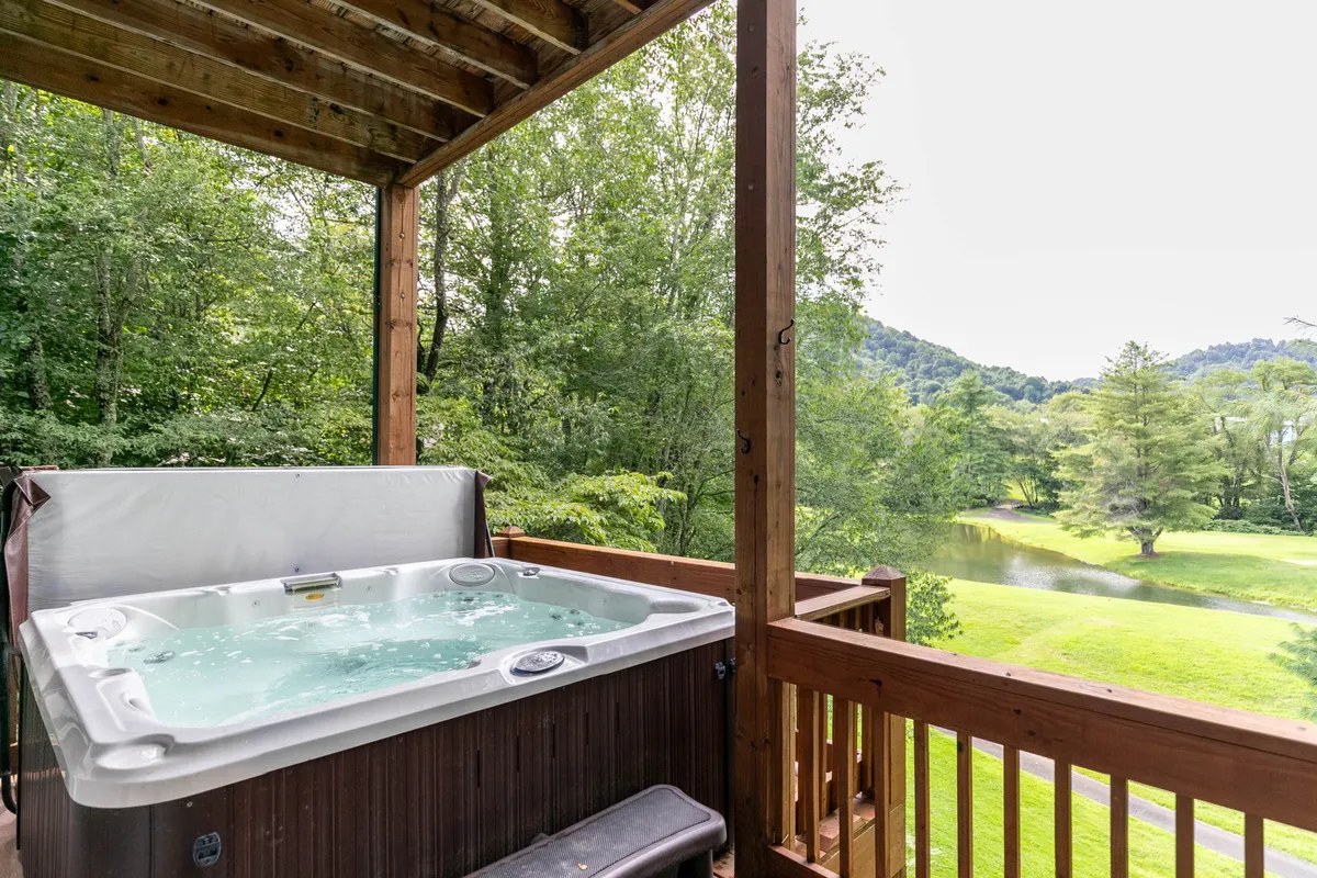 The image showcases the rustic wooden deck of A Happy Roost cabin, with a steaming hot tub perfectly positioned to overlook a lush natural landscape. In the distance, you can see the garden-like golf course, a tranquil pond, and layers of blue mountain ridges fading into the sunset. The scene suggests comfort, relaxation, and an immersive experience in nature, inviting guests to unwind outdoors.